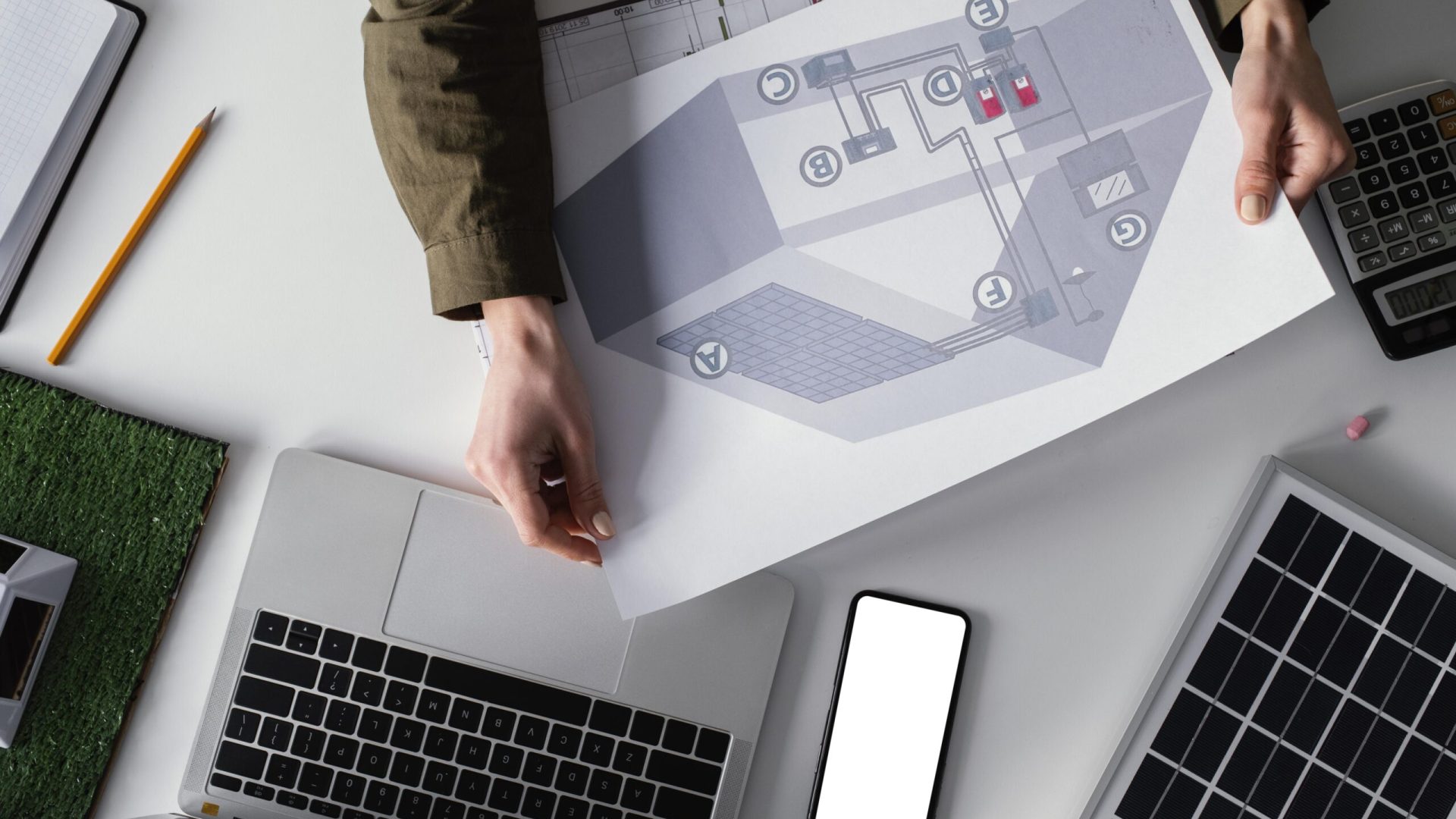 A woman holds a paper with a house illustration and points inside, next to a laptop, suggesting property management software use.